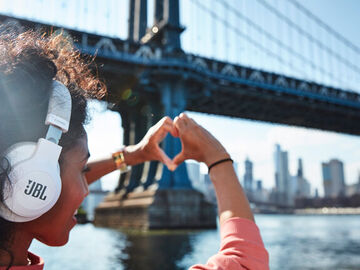 A woman with JBL headphones forms a heart with her fingers and points it towards Manhattan