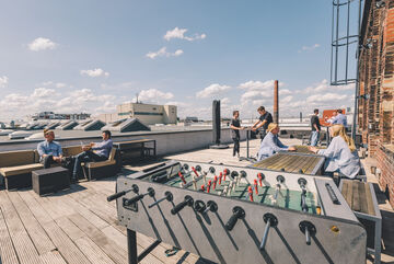 eine Dachterrasse unter blauem Himmel mit einem Kickertisch im Vordergrund und mehreren Mitarbeitern, die sich im Hintergrund unterhalten